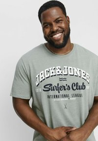 Zaļš kokvilnas t-krekls ar īsām piedurknēm, uz kura priekšpuses ir balta un jūraszilā teksts: "JACK & JONES Surfer's Club" un grafika.