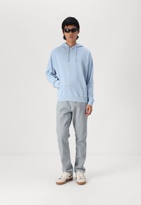 Felpa azzurra con tasca a marsupio e cordino, pantaloni grigi e sneakers bianche con suola marrone. Il modello indossa occhiali da sole neri.