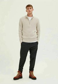 Bežinis raštuotas megztinis su užtrauktuku prie apykaklės, derinamas su tamsiais dryžuotais kelniais ir rudais bateliais su šviesiomis raištelėmis.