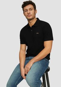 Schwarzes Poloshirt mit einem Kragen, versehen mit einem goldenen Logo auf der Brust, kombiniert mit hellblauen Jeans. Modell sitzt auf einem schwarzen Hocker.