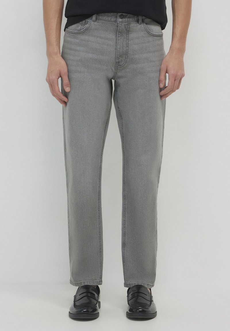 Homme portant un jean gris droit et des mocassins en cuir noir, se tenant devant un fond blanc uni.