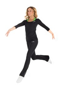 Schwarzes Sportoberteil mit drei Viertelärmeln, kombiniert mit schwarzen, enganliegenden Hosen. Auffälliger grüner Akzent auf dem Oberteil. Modell trägt weiße Turnschuhe.