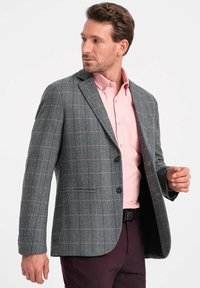 Blazer grigio a quadri con accenti neri, caratterizzato da una vestibilità slim, rever a punta, due bottoni e una texture morbida. Indossato sopra una camicia rosa.