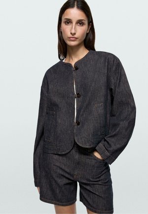 Jeune femme portant une veste en jean foncé boutonnée avec poches plaquées et un short assorti, debout avec une main dans la poche sur un fond clair.