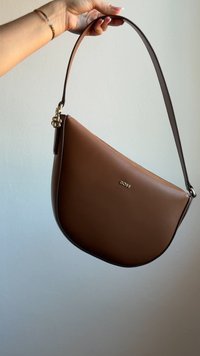 Main tenant un sac bandoulière BOSS marron à la forme incurvée et à la quincaillerie dorée, présenté sur un fond clair uni.