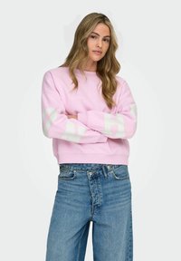 Helehelenroosa dressipluus valgete plokktähtedega varrukatel. Omaduseks on ümmargune kaelus ja lühendatud ääreosa. Kantud siniste kõrge vöökohaga teksadega.