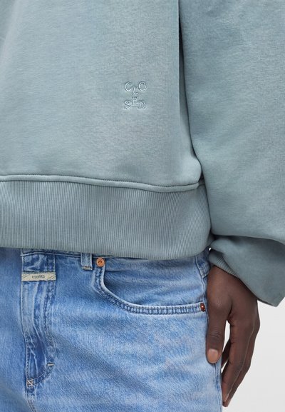 Sweat-shirt bleu clair avec petit texte brodé "CLOSED" porté sur un jean denim bleu clair avec une étiquette "CLOSED" assortie près de la poche.