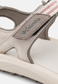 Sandalia beige con un upper de ante texturizado y un forro de malla oscura. Incluye un logo en color azul verdoso, acentos de tiras a rayas y una suela de goma.