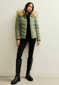 Vrouw met een groene gevoerde jas met een capuchon van bont, zwarte legging en hoge zwarte laarzen, staand tegen een lichte muur.