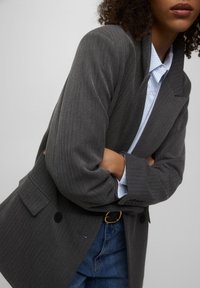 Veste grise à fines rayures avec une coupe structurée, des revers crantés et des poches à rabat. Portée sur une chemise à rayures bleu clair, associée à un jean bleu.