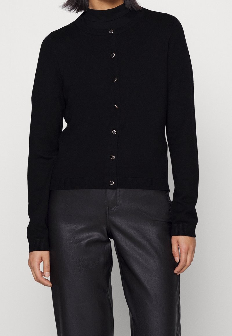 Femme portant un cardigan noir à manches longues avec de petits boutons en forme de cœur et un pantalon noir en simili cuir, debout devant un fond blanc.