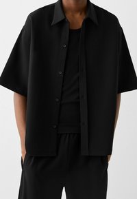Schwarzes kurzärmeliges Hemd aus leichtem Material, mit Knopfverschluss an der Vorderseite und einer lockeren Passform. Tragend über einem passenden schwarzen Tanktop.