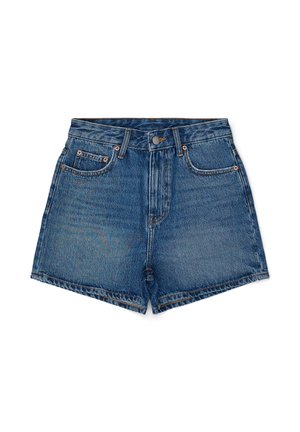 Ung kvinde, der står med front mod kameraet iført en hvid tanktop, denimshorts, mørke strømper, sorte sneakers og to halskæder på en ensartet lys baggrund.
