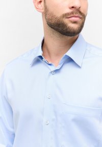 Camicia azzurra chiara con bottoni, colletto strutturato, taschino frontale e sottile texture a righe diagonali. I bottoni sono bianchi.