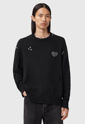 Uomo con capelli lunghi che indossa un maglione nero strappato con una patch a forma di cuore sul petto, in piedi con una mano in tasca su sfondo grigio.