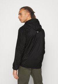 Black lightweight jacket with a hood, textured fabric, and a discreet logo on the back. Features long sleeves and a relaxed fit.