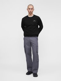 Homme à la tête rasée portant un sweat-shirt noir avec un logo et un pantalon cargo gris, debout devant un fond blanc uni.