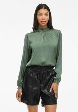 Vrouw in een lange mouwen groene satijnen blouse, erin gestoken in een hoge taille zwarte leren shorts met elastische tailleband.