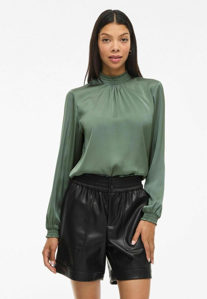 Femme portant un chemisier en satin vert à manches longues rentré dans un short en cuir noir taille haute avec une ceinture élastique.