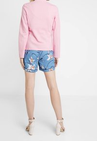 Osoba v růžovém tričku s dlouhým rukávem, modrých květinových kraťasech a béžových vysokých podpatcích, stojící zády k bílé pozadí.