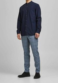 Camicia blu navy con colletto a bandana, abbinata a pantaloni a quadri blu e grigi, completata da scarpe nere, in piedi su uno sfondo chiaro.