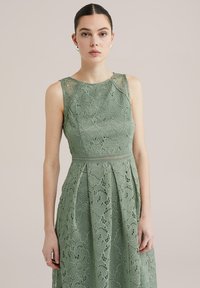 Spetsklänning i mjukt grönt, med rund halsringning, ärmlös design och en figurnära midja med plisserad kjol. Utsmyckad med blommönster.
