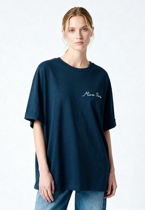 Jonge vrouw met blond haar draagt een oversized donkerblauw T-shirt met de tekst "Mira Paris" en lichtblauwe jeans, staand tegen een witte achtergrond.