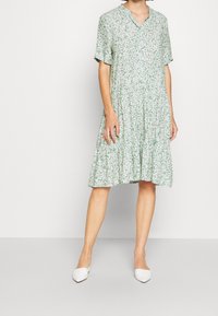 Abito floreale verde con maniche corte, scollatura a V e gonna a strati. Presenta un motivo di margherite bianche su un tessuto leggero. Abbinato a scarpe bianche.