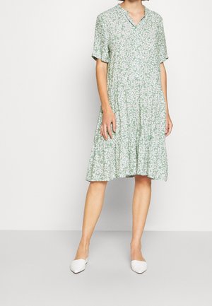 Robe florale verte à manches courtes, décolleté en V et jupe à volants. Présente un motif de marguerites blanches sur un tissu léger. Associée à des chaussures blanches.