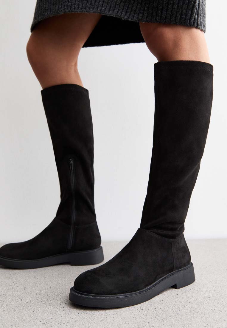 Schwarze kniehohe Wildlederstiefel mit flachen Sohlen, getragen zu einem dunkelgrauen gerippten Strickrock, auf einem hellgrauen Boden und vor einem weißen Hintergrund gezeigt.