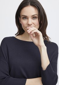 Jūras spēku zili ribota adījuma džemperis ar plašu kuģa kakla izgriezumu un trim ceturtdaļām garām piedurknēm. Modelis viegli balstījis roku uz zoda, parādot mīksto tekstūru.