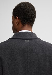 Donkergrijze wollen jas met een brede kraag, voorzien van een klein label op de achterkant en een gestructureerd oppervlak. Zichtbare stiksels langs de naden.