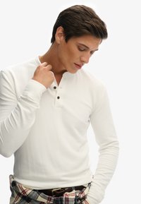 Chemise henley blanche à manches longues avec trois boutons marron au cou, présentant un design ajusté et une texture lisse associée à un pantalon à carreaux.