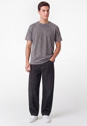 Giovane uomo in piedi con una mano in tasca che indossa una t-shirt grigia lavata, jeans larghi e scuri e sneakers bianche su sfondo bianco.