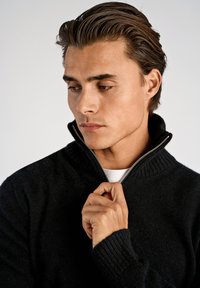 Schwarzer, strukturierter Pullover mit hohem Kragen und Reißverschluss-Detail; Modell hält den Reißverschluss, Nahaufnahme, hellgrauer Hintergrund.