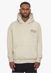 Beige hettegenser med frontlomme, med en liten brun logo på venstre side. Laget av myk stoff med en avslappet passform.