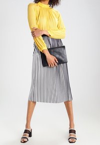 Haut à manches longues texturé jaune associé à une jupe midi rayée noir et blanc, tenant une pochette noire. Des sandales noires à talons complètent le look.