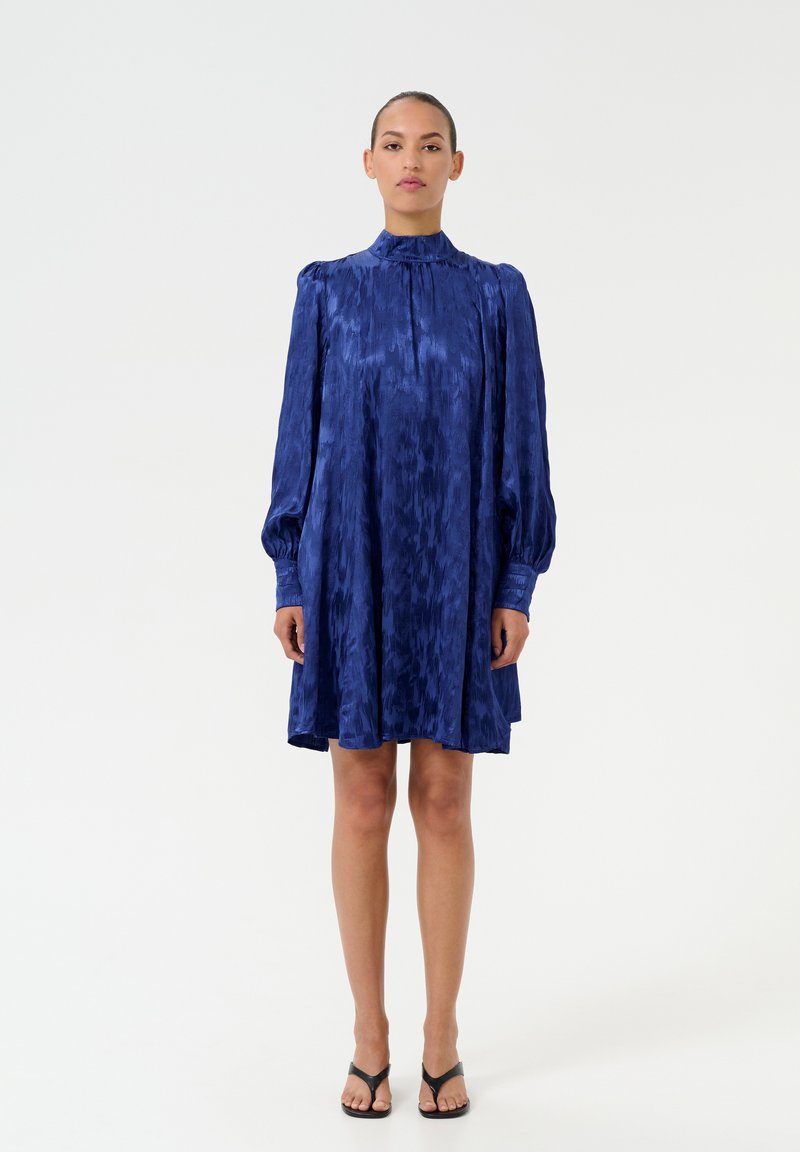 Rochie din satin albastru cu gât înalt, mâneci lungi în puff și croială lejeră. Prezintă o textură subtilă și se termină deasupra genunchilor.
