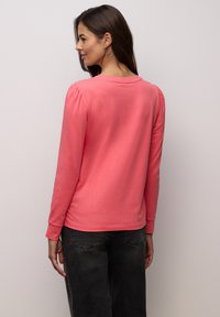 Långärmad top i korallfärgad bomull med puffade axlar, rundad halsringning och ribbade muddar; kombinerad med mörkgrå byxor.