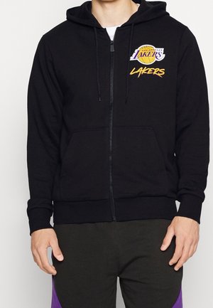 Osoba nosząca czarną bluzę z kapturem na zamek z logo Los Angeles Lakers i tekstem na lewej piersi, w połączeniu z czarnymi i fioletowymi spodniami.