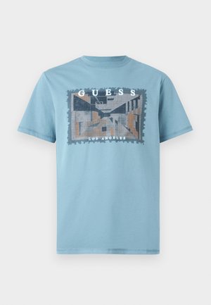Camiseta de algodón azul claro con un diseño gráfico rectangular que presenta patrones geométricos y el texto "GUESS LOS ÁNGELES". Mangas cortas y cuello redondo.