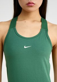 Žalias sportinis tankinis su apvaliu kaklu, storomis petnešomis ir mažu baltu Nike logotipu ant krūtinės. Medžiaga atrodo lygi ir elastinga.