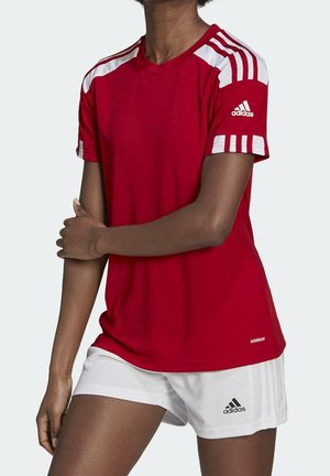 Osoba nosząca czerwoną koszulkę Adidas z krótkim rękawem z białymi paskami oraz białe szorty Adidas, stojąca z jedną ręką skrzyżowaną nad drugą.