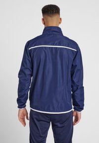 Marinblå vindjacka med hög krage, prydd med vita horisontella detaljer över axlarna och ryggen. Lättviktig, slät textur.