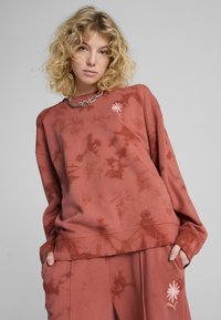 Pulover v rožnato barvi z potiskom tie-dye, s vezenim cvetličnim detajlom in rebrastimi manšetami, v kombinaciji z usklajenimi hlačami, kar ustvarja sproščen silhuet.