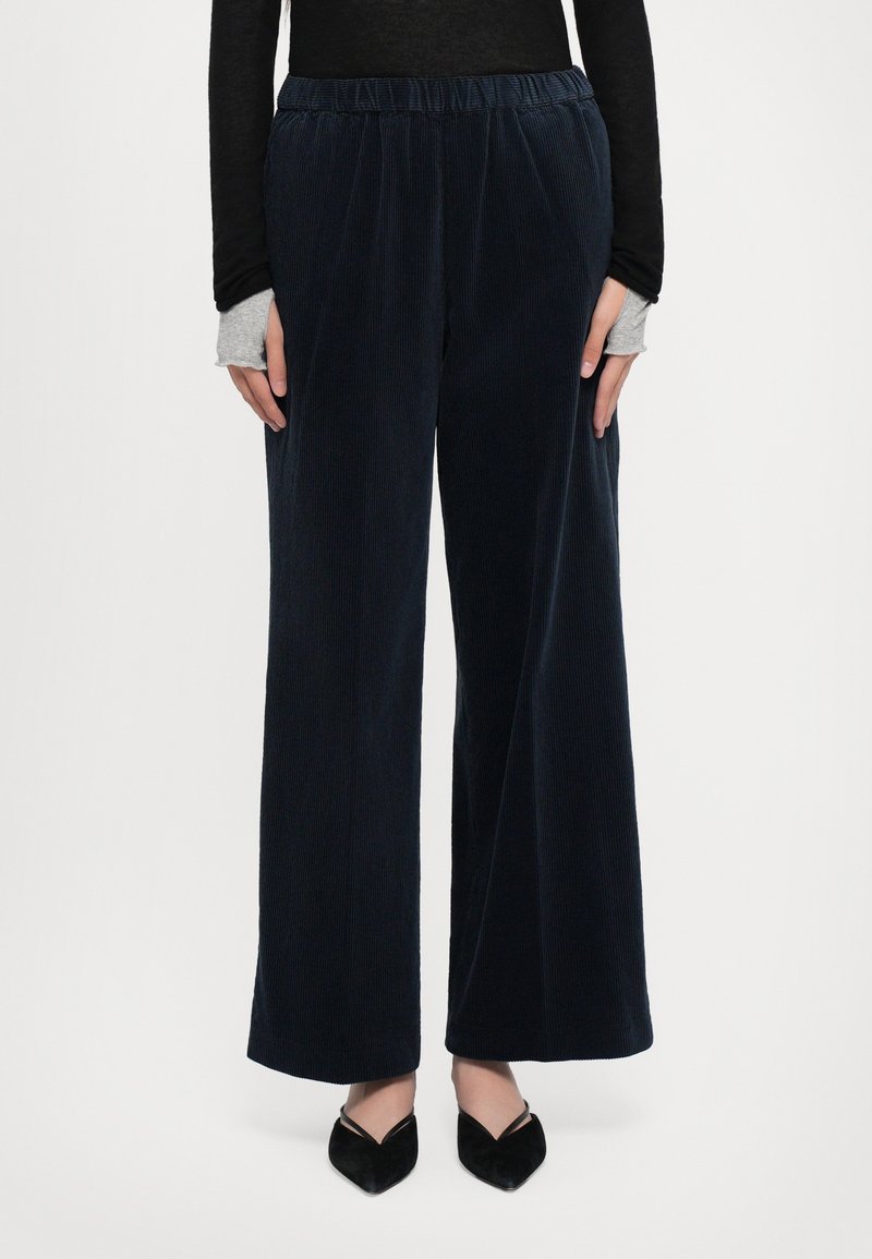 Femme portant un pantalon large en velours côtelé bleu marine avec une taille élastique, des chaussures noires pointues et des hauts à manches longues superposés avec des trous pour les pouces.