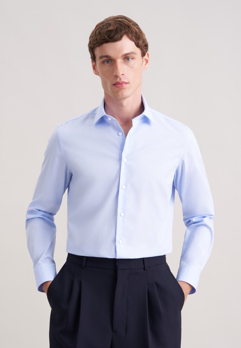 Un jeune homme aux cheveux châtain clair porte une chemise habillée bleu clair boutonnée et un pantalon noir plissé, debout les mains dans les poches.