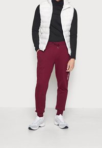 Weiße, puffige Steppweste über einem schwarzen Langarmshirt; dunkelrote Jogginghosen mit Kordelzug; weiße Turnschuhe mit grauen Akzenten.