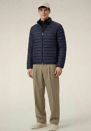 Giovane uomo con giacca trapuntata blu navy, pantaloni beige, cappellino beige e scarpe da ginnastica beige, in piedi davanti a uno sfondo semplice.