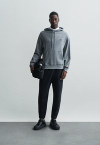 Sivina kapuca z vrvicami in sprednjim žepom, v kombinaciji s črnimi ožjimi hlačami in črnimi supergami. Model drži črn nahrbtnik.